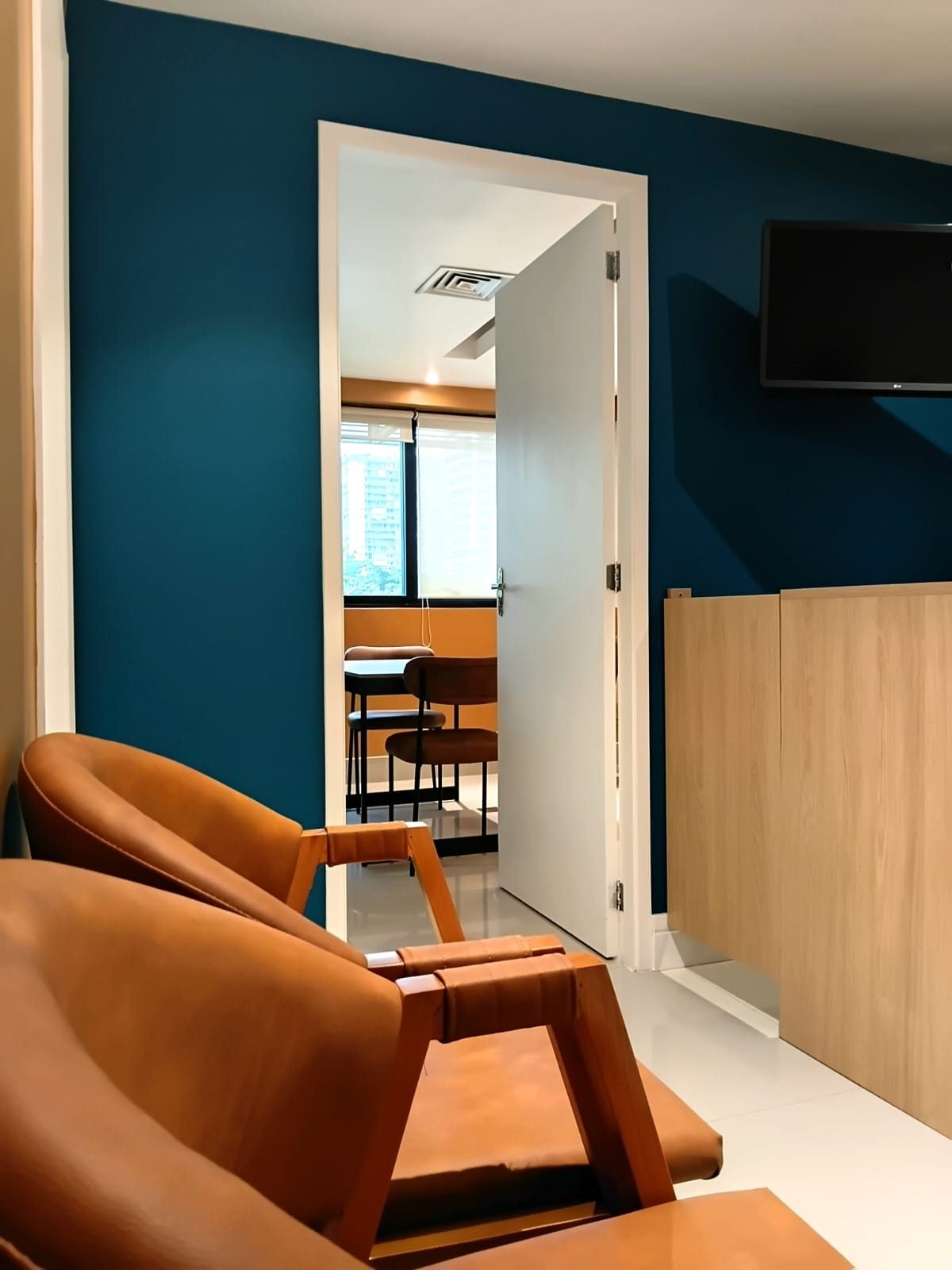 Área de espera da clínica com poltronas de couro caramelo, parede verde-azulada e porta do consultório ao fundo.